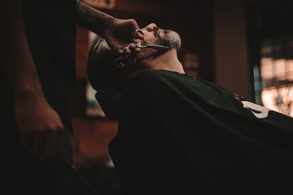 Barber trimming a client's beard with shears in a low-lit shop